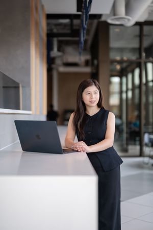 Professional Asian woman in black outfit leaning on white desk with laptop in modern office.