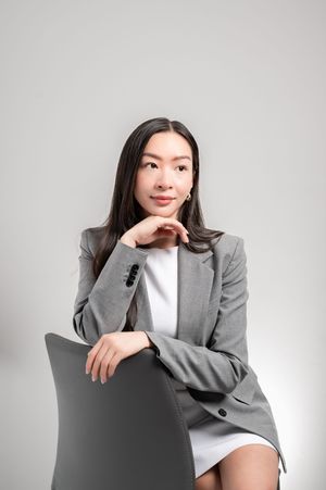 Young Asian businesswoman in gray blazer sitting confidently on chair with hand on chin against white background.