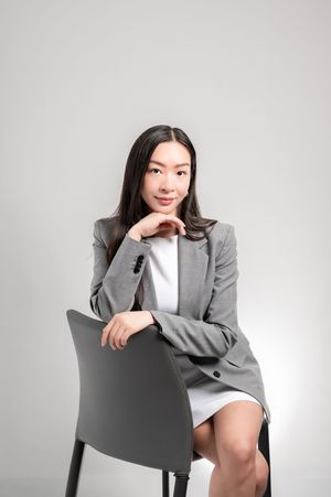 Young Asian businesswoman in gray blazer sitting confidently on chair with hand on chin, studio portrait.