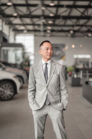 A professional man in a light gray suit stands confidently in a modern car dealership showroom.