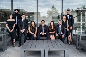 Group of nine professionals in formal black attire posing outdoors on a modern patio for a team photo.