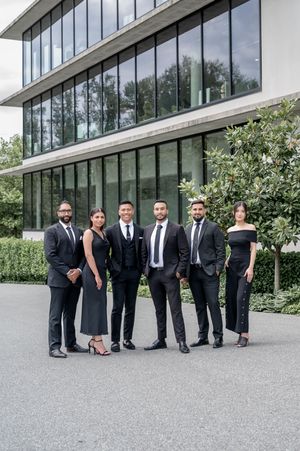 Six professionals in black formal attire posing together outside a modern glass office building.