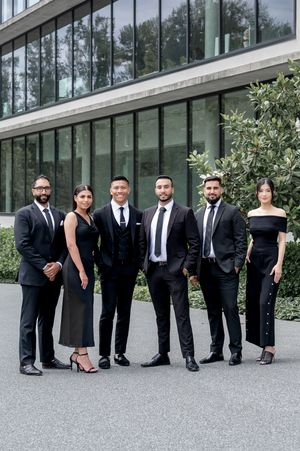 Six professionals in black formal attire posing outdoors in front of a modern glass office building.