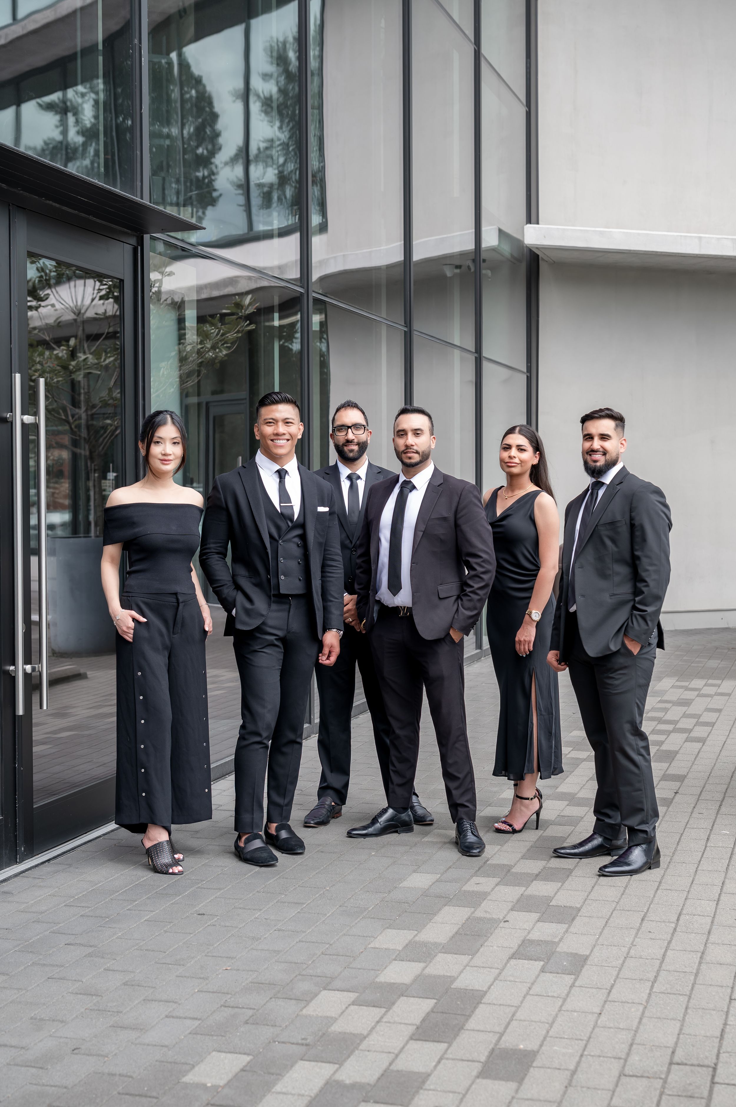 Six professionals in elegant black attire pose confidently outside a modern glass office building.