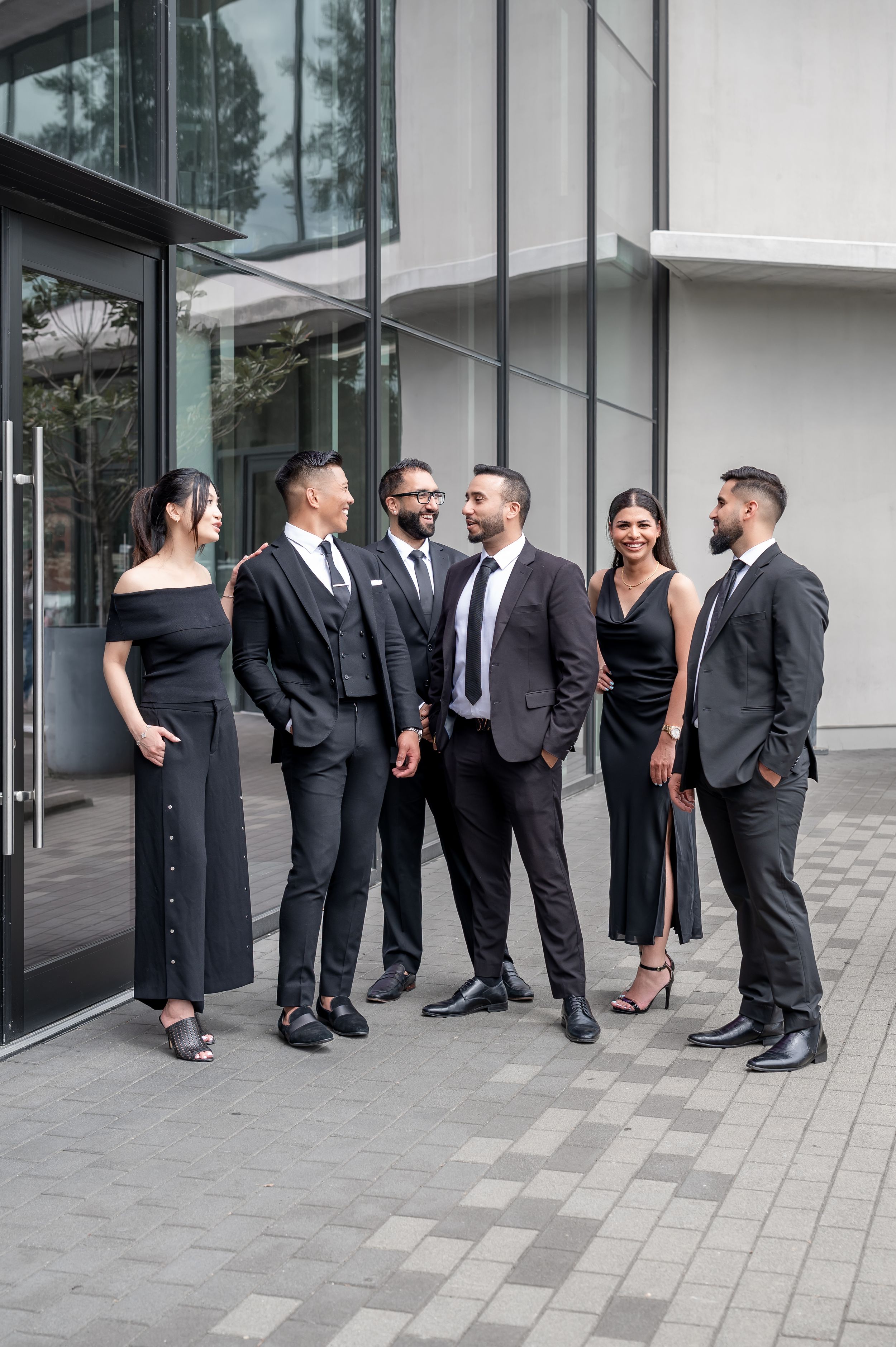 Six elegantly dressed people in black formal attire posing together outside a modern glass building.