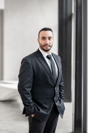 Professional man in black suit and tie standing confidently indoors, suitable for business or corporate profile.