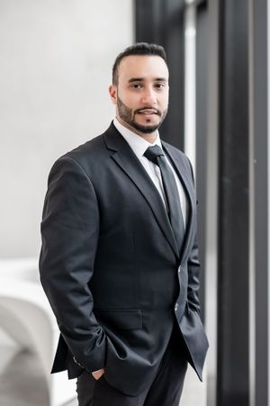 Professional businessman in black suit and tie with beard standing confidently in modern office setting.