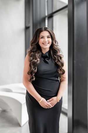 Professional woman in black sleeveless dress with long wavy hair smiling confidently in modern interior setting.