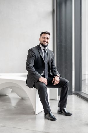 Smiling bearded businessman in dark suit sitting on white bench in modern office lobby.
