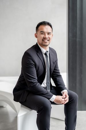 A professional Asian man in a black suit and tie sits smiling confidently in a modern minimalist setting.