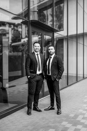 Two professional businessmen in black suits and ties standing outside a modern glass office building, black and white photo.