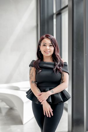Woman in black peplum top and leggings with tattoos, posing confidently in a modern bright interior space.