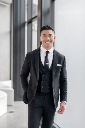 Smiling young Asian businessman in a dark three-piece suit with white shirt and tie standing in modern office.