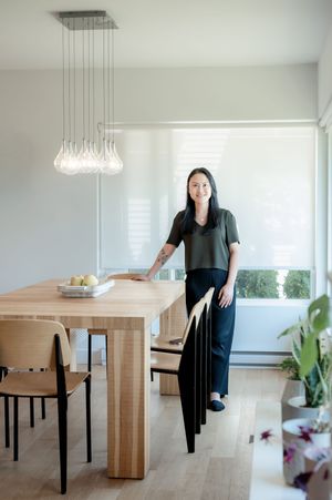 Woman in dark outfit stands beside a light wood dining table under modern glass pendant lights in a bright minimalist room.