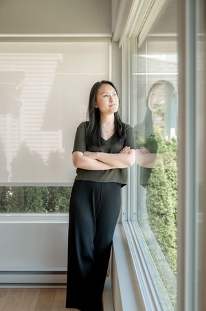 A woman in a green top and black pants stands with arms crossed by a large window, gazing thoughtfully outside.