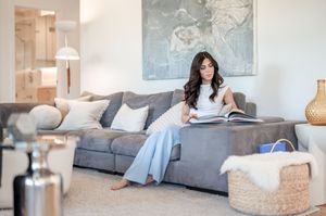 Woman in white top and light blue pants reading a magazine on a gray sofa in a modern living room.