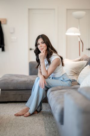 Woman in white top and light blue pants sitting relaxed on a gray sofa in a modern living room.