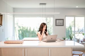 Woman standing at a modern kitchen island, covering her mouth while working on a laptop computer.
