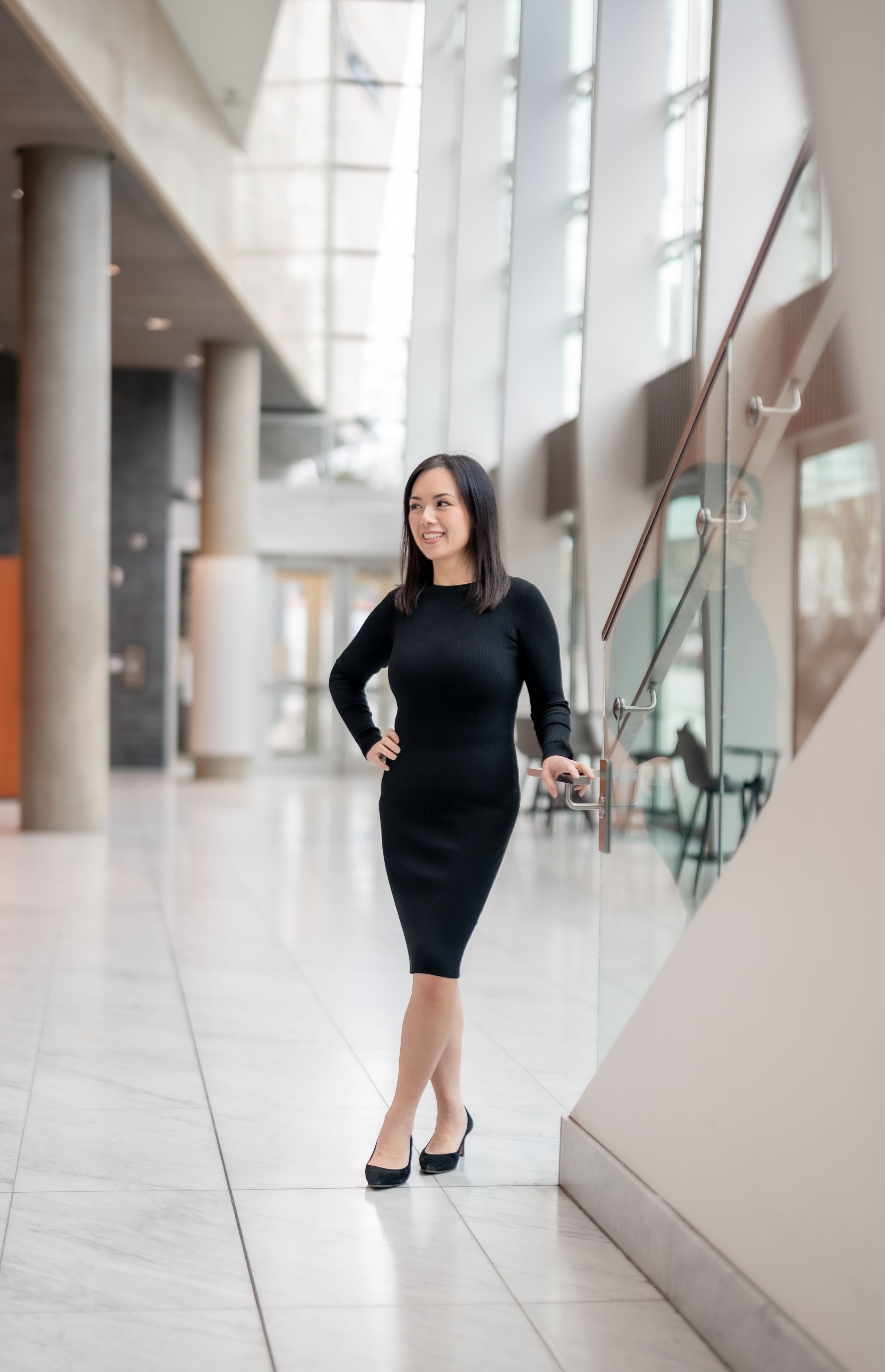 A professional in a fitted black dress and heels poses confidently in a modern office building with large windows.