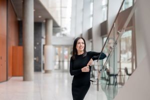 Professional in black attire poses confidently against modern office interior with glass railings and columns.