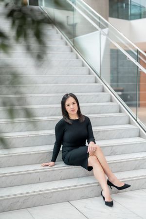 Woman in black dress and heels sitting elegantly on modern concrete stairs with glass railing.