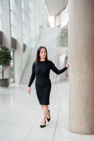 Professional figure in a fitted black dress and heels poses elegantly in a modern bright interior with columns.