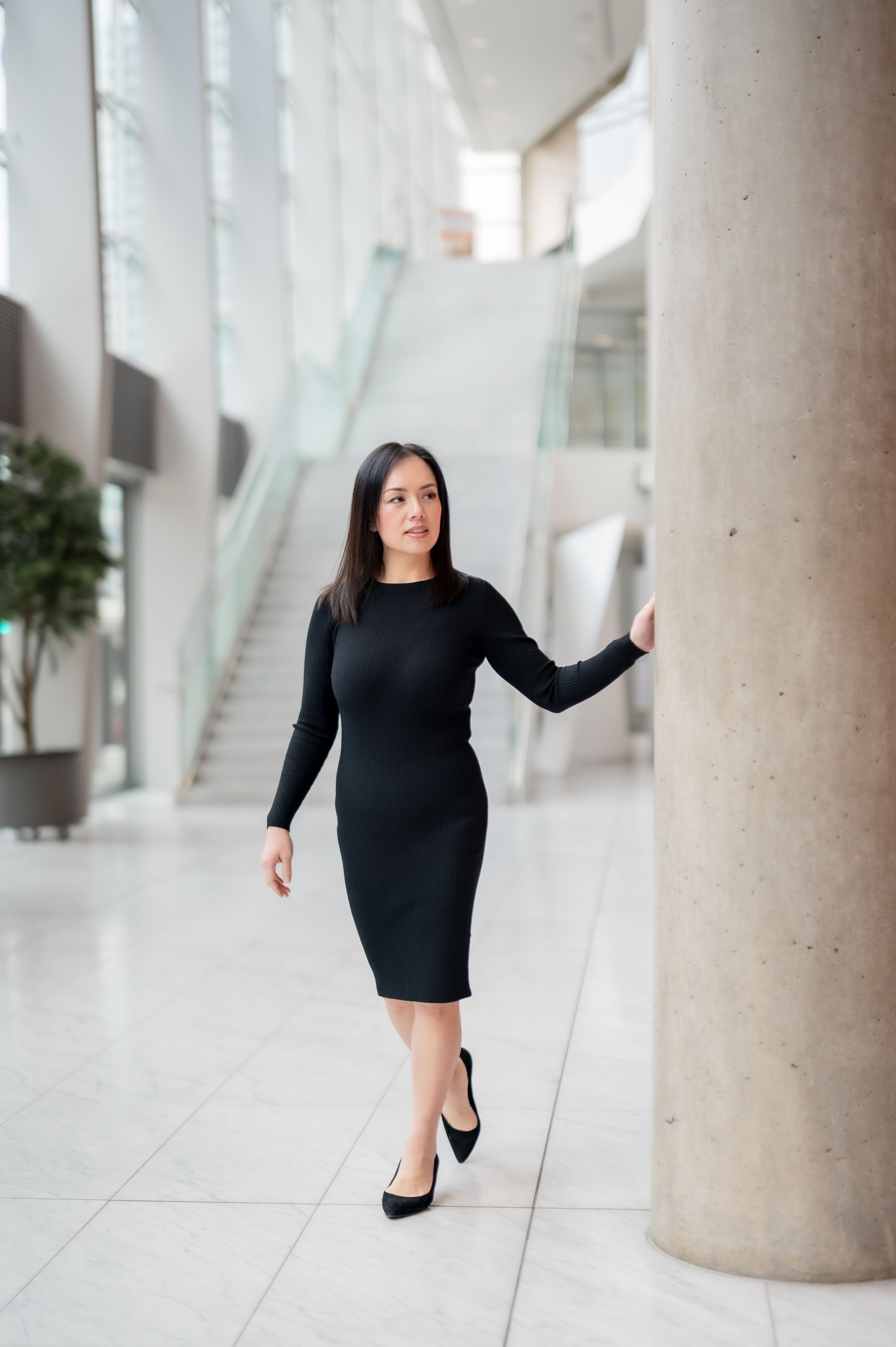 Professional figure in a fitted black dress and heels poses elegantly in a modern bright interior with columns.