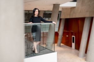 A person wearing a black dress stands near a glass railing in a modern office building interior.