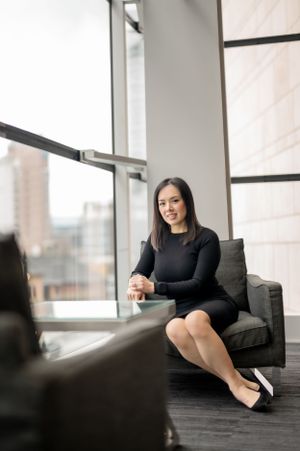 A woman in a black dress sits in a modern office chair by large windows while engaged in a professional conversation.