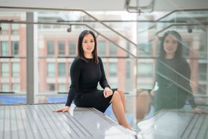 Professional in black sweater sits gracefully on modern staircase with glass railings and natural lighting.