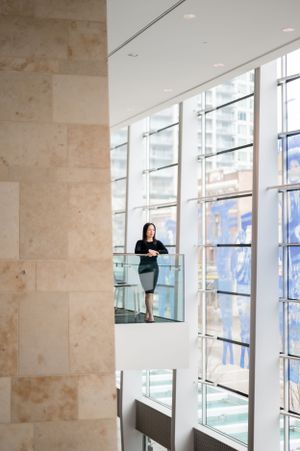 Person stands on modern glass walkway overlooking bright atrium with tall windows and beige stone walls.