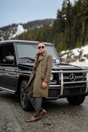 A black Mercedes-Benz G-Wagon parked on a snowy mountain road with a stylishly dressed person leaning against it.
