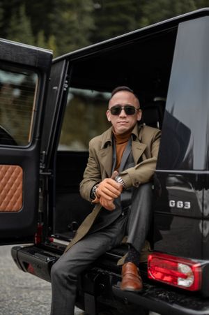Stylishly dressed person in sunglasses and tan coat sits casually in back of luxury SUV wearing brown leather shoes.