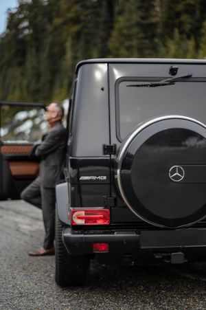 Rear view of a black Mercedes-AMG G-Class SUV parked on a wet road with trees in the background.