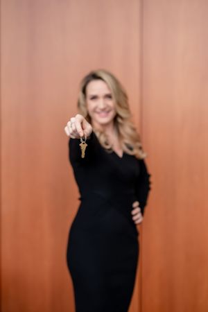 Woman in elegant black dress holds up keys while posing against terra cotta colored wall background.