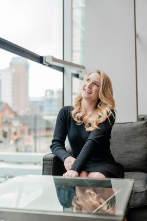 Woman in black sweater dress sitting on grey couch near window with cityscape view.