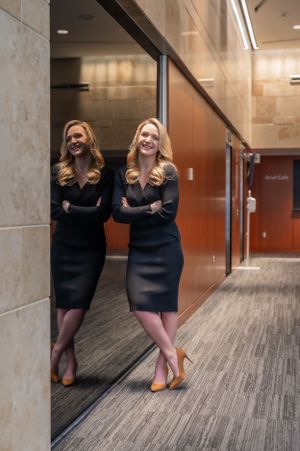 Professional in a black dress standing next to a mirror in an office hallway with carpeted floors.
