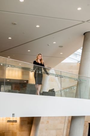 A person in a black dress stands at a modern glass balcony railing in a corporate building interior with recessed lighting.