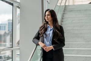 Professional in a black blazer and blue button-up shirt stands thoughtfully by a modern glass window with stairs.