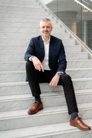 A professional in a navy blazer and white shirt sits confidently on modern concrete stairs wearing brown leather shoes.