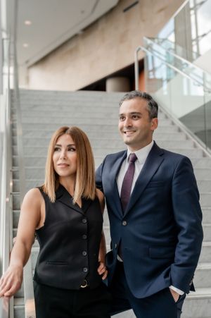 Two professionals in business attire stand together in a modern office building with glass stairs and neutral decor.