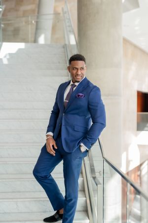 Professional wearing a tailored navy blue suit and burgundy tie poses confidently on marble stairs in modern building.