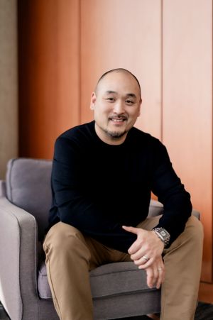 Casual professional in black sweater and khaki pants sits relaxed in gray armchair against orange wall.