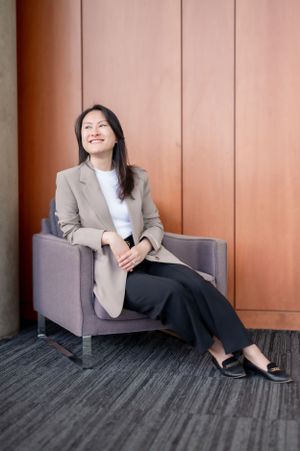 Professional wearing a beige blazer and white top sits relaxed in a gray armchair against an orange wall.