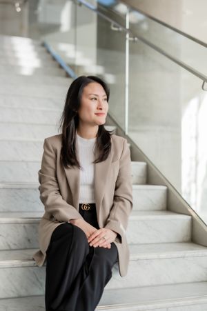 A person in a beige blazer and white sweater sits thoughtfully on marble stairs in a modern office building.
