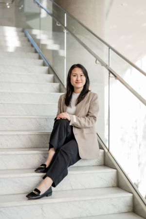 Professional woman in beige blazer and dark pants sits casually on white marble stairs in modern interior setting.