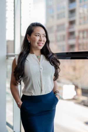 Professional woman in a white button-down blouse and navy pencil skirt poses confidently by bright office window.