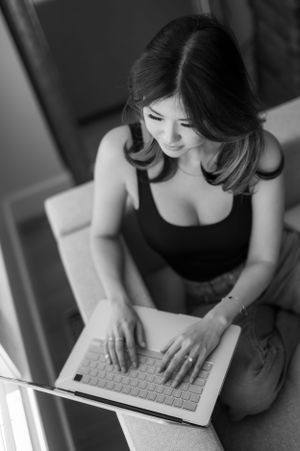 Black and white photo of woman in dark top typing on laptop keyboard while sitting on floor near window.