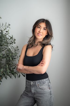 A woman in a black tank top and gray jeans poses casually against a light gray wall with a potted plant nearby.