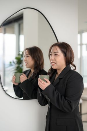 A person in a black blazer stands beside a round wall mirror holding a cup in a bright interior space.